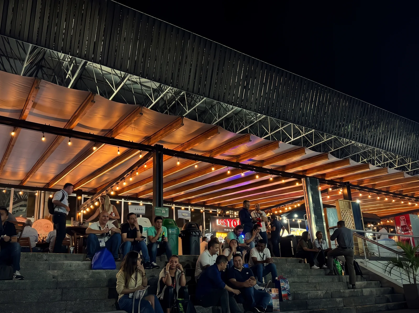 Pérgola de madera iluminada de noche con guirnaldas de bombillos cálidos sobre el público